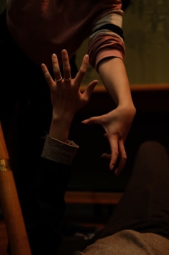Two hands reaching towards each other in a dimly lit environment, with a focus on the expressive gesture of the fingers. The setting suggests a moment of connection or support, with clothing indicating casual attire.