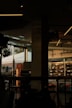 The image features a dimly lit café interior with modern design elements. On the right side, a glass refrigerator displays organic products. The left side of the image shows large windows that let in natural light, showcasing a view of people walking outside and a prominent sign with the word 'La Gamba'. The interior features dark furniture and ceiling lights with a minimalist style.