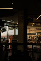 The image features a dimly lit caf&eacute; interior with modern design elements. On the right side, a glass refrigerator displays organic products. The left side of the image shows large windows that let in natural light, showcasing a view of people walking outside and a prominent sign with the word 'La Gamba'. The interior features dark furniture and ceiling lights with a minimalist style.