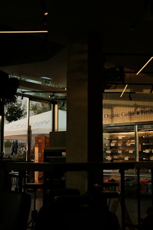 The image features a dimly lit caf&eacute; interior with modern design elements. On the right side, a glass refrigerator displays organic products. The left side of the image shows large windows that let in natural light, showcasing a view of people walking outside and a prominent sign with the word 'La Gamba'. The interior features dark furniture and ceiling lights with a minimalist style.