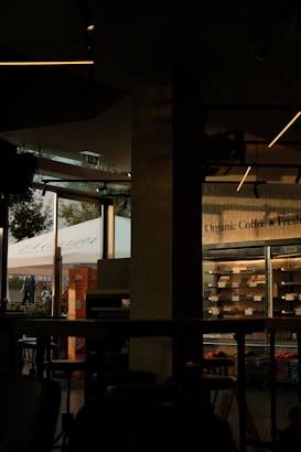 The image features a dimly lit café interior with modern design elements. On the right side, a glass refrigerator displays organic products. The left side of the image shows large windows that let in natural light, showcasing a view of people walking outside and a prominent sign with the word 'La Gamba'. The interior features dark furniture and ceiling lights with a minimalist style.