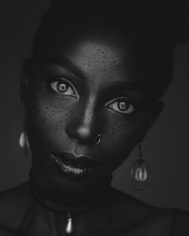 A grayscale portrait of a person with expressive eyes looking directly at the viewer. The skin is speckled with a texture or freckles, and the person is adorned with a nose ring and unique earrings featuring circular and teardrop shapes. The image focuses on facial features and expressions with a smooth and detailed texture.