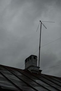 A rooftop with a weathered chimney and an old television antenna against a backdrop of overcast gray clouds, suggesting an impending storm or rain.