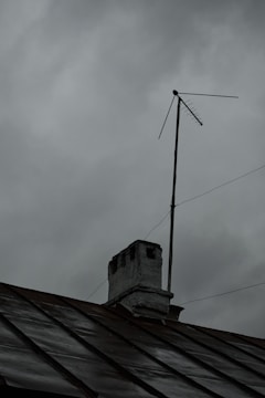 A rooftop with a weathered chimney and an old television antenna against a backdrop of overcast gray clouds, suggesting an impending storm or rain.