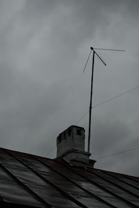 A rooftop with a weathered chimney and an old television antenna against a backdrop of overcast gray clouds, suggesting an impending storm or rain.
