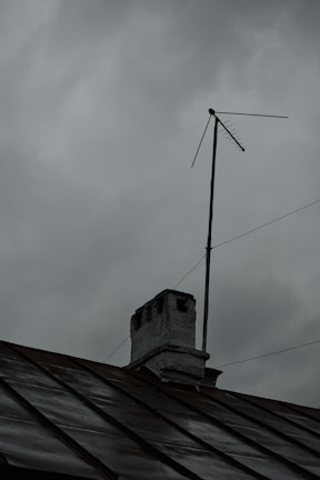 A rooftop with a weathered chimney and an old television antenna against a backdrop of overcast gray clouds, suggesting an impending storm or rain.