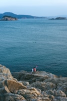 A couple sits on rocky cliffs overlooking the calm sea. The distant view includes several small islands and a serene horizon. The couple is holding a selfie stick, capturing the moment.