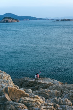 A couple sits on rocky cliffs overlooking the calm sea. The distant view includes several small islands and a serene horizon. The couple is holding a selfie stick, capturing the moment.