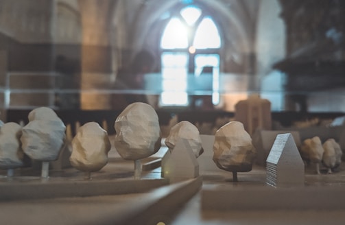 A miniature architectural model showcases abstract, geometric trees and buildings under soft lighting. The setting appears to be indoors, with an arched window in the background allowing natural light to filter through, creating a calm and contemplative atmosphere.