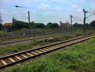 Railway tracks crossing through a green field, accompanied by overhead electrical wires supported by metal poles. In the background, there are piles of gravel, trees, and a few buildings under a partly cloudy sky.