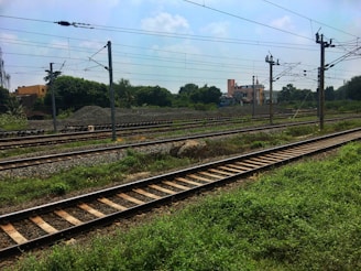 Railway tracks crossing through a green field, accompanied by overhead electrical wires supported by metal poles. In the background, there are piles of gravel, trees, and a few buildings under a partly cloudy sky.