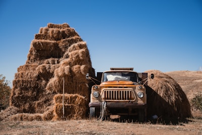 An old, rusted truck is parked next to a large stack of hay bales under a clear blue sky. The bales are neatly arranged into a pyramid shape, with some scattered around the truck. The landscape is dry and open, giving a deserted and rustic feel.