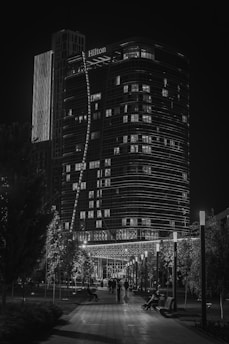 A nighttime cityscape featuring a tall, illuminated hotel building with a modern design, surrounded by trees and a lit pathway. People are walking along the walkway, and decorative lights adorn the pathway on either side, creating a serene and urban atmosphere.