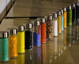 A series of glass jars filled with colored powders and granules arranged in a line on a smooth surface. Each jar is topped with a metallic lid, and the colors range from green, yellow, black, blue, to red. The image features a reflective shine on the surface adding depth to the arrangement.