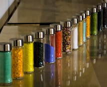 A series of glass jars filled with colored powders and granules arranged in a line on a smooth surface. Each jar is topped with a metallic lid, and the colors range from green, yellow, black, blue, to red. The image features a reflective shine on the surface adding depth to the arrangement.