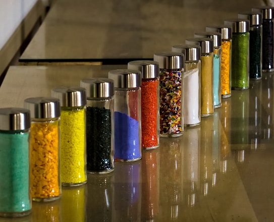 A series of glass jars filled with colored powders and granules arranged in a line on a smooth surface. Each jar is topped with a metallic lid, and the colors range from green, yellow, black, blue, to red. The image features a reflective shine on the surface adding depth to the arrangement.