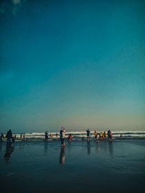 A group of people stands on a sandy beach near the ocean, with waves gently rolling in the background. Some individuals are wearing colorful clothing and are either wading into the water or standing by the shore. The sky above is clear with a gradient of blue.