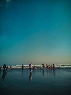 A group of people stands on a sandy beach near the ocean, with waves gently rolling in the background. Some individuals are wearing colorful clothing and are either wading into the water or standing by the shore. The sky above is clear with a gradient of blue.