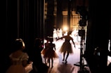 Several dancers, possibly in costumes, are gathered backstage, preparing to perform. The light from the stage creates a silhouette effect on the dancers, highlighting their movements. The stage is illuminated, and more performers can be seen in the distance, already engaged in a dance routine.
