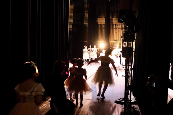 Several dancers, possibly in costumes, are gathered backstage, preparing to perform. The light from the stage creates a silhouette effect on the dancers, highlighting their movements. The stage is illuminated, and more performers can be seen in the distance, already engaged in a dance routine.