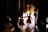 Several dancers, possibly in costumes, are gathered backstage, preparing to perform. The light from the stage creates a silhouette effect on the dancers, highlighting their movements. The stage is illuminated, and more performers can be seen in the distance, already engaged in a dance routine.