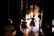 Several dancers, possibly in costumes, are gathered backstage, preparing to perform. The light from the stage creates a silhouette effect on the dancers, highlighting their movements. The stage is illuminated, and more performers can be seen in the distance, already engaged in a dance routine.