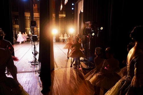 Ballet dancers in elegant costumes prepare backstage under the warm glow of stage lights, with some performers already on stage in graceful poses.