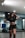 A woman with long dark hair stands confidently in a gym or dance studio. She is wearing a black crop top and dark shorts, posing with one hand on her hip and the other touching her hair. The room has mirrors, a hardwood floor, and equipment like a blue mat in the background.