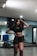 A woman with long dark hair stands confidently in a gym or dance studio. She is wearing a black crop top and dark shorts, posing with one hand on her hip and the other touching her hair. The room has mirrors, a hardwood floor, and equipment like a blue mat in the background.