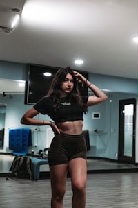 A woman with long dark hair stands confidently in a gym or dance studio. She is wearing a black crop top and dark shorts, posing with one hand on her hip and the other touching her hair. The room has mirrors, a hardwood floor, and equipment like a blue mat in the background.