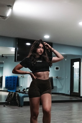 A woman with long dark hair stands confidently in a gym or dance studio. She is wearing a black crop top and dark shorts, posing with one hand on her hip and the other touching her hair. The room has mirrors, a hardwood floor, and equipment like a blue mat in the background.