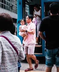 A man is carrying a young child as they step off a blue public transportation vehicle. The child looks cheerful, and several other people are around, with one person preparing to step off the vehicle while looking at a phone. The scene is bustling with activity and urban life.
