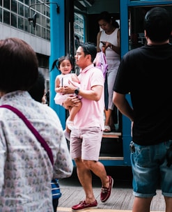 A man is carrying a young child as they step off a blue public transportation vehicle. The child looks cheerful, and several other people are around, with one person preparing to step off the vehicle while looking at a phone. The scene is bustling with activity and urban life.