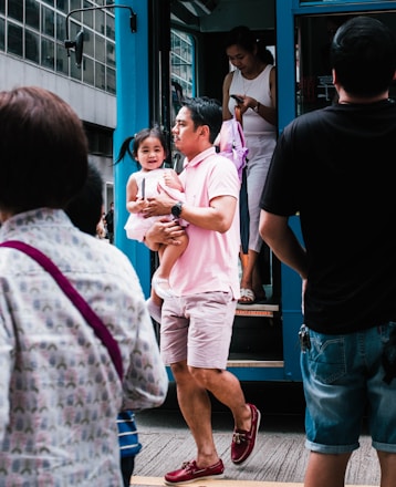 A man is carrying a young child as they step off a blue public transportation vehicle. The child looks cheerful, and several other people are around, with one person preparing to step off the vehicle while looking at a phone. The scene is bustling with activity and urban life.
