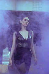 A woman stands surrounded by a mystical purple haze, wearing a black outfit with dark wings. The atmosphere feels ethereal and dramatic, enhanced by the fog and subdued lighting.