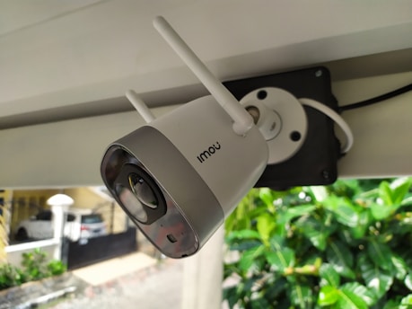 A security camera with two antennas is mounted on a structure. The camera is branded with the name 'Imou' and is connected to a black mounting plate. Background includes a parked car, a building, and green foliage.