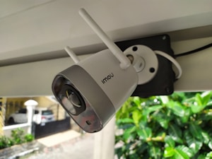 A security camera with two antennas is mounted on a structure. The camera is branded with the name 'Imou' and is connected to a black mounting plate. Background includes a parked car, a building, and green foliage.