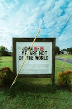 A wooden sign stands on a grassy area next to a road, displaying the message 'JOHN 15:19 YE ARE NOT OF THE WORLD' in bold black and red letters. A yellow pole is leaning against the sign. The sky is partly cloudy, and there are trees and shrubs in the background.