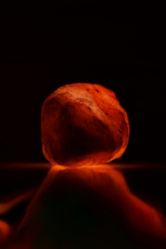 A glowing rock with a warm, orange hue, possibly made of Himalayan salt, resting on a reflective surface. The background is dark, emphasizing the illumination of the rock.