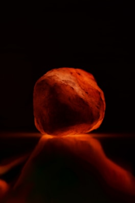 A glowing rock with a warm, orange hue, possibly made of Himalayan salt, resting on a reflective surface. The background is dark, emphasizing the illumination of the rock.