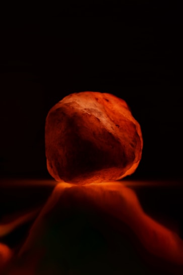 A glowing rock with a warm, orange hue, possibly made of Himalayan salt, resting on a reflective surface. The background is dark, emphasizing the illumination of the rock.