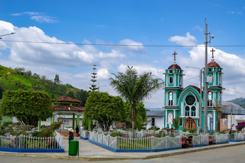 A picturesque scene featuring a small town plaza with a vibrant turquoise church, adorned with two bell towers capped with brown domes and crosses. It is surrounded by well-maintained gardens with various trees and flowers, and white railings outlining pathways. A mountainous backdrop with lush greenery and a partly cloudy sky adds to the scenic beauty. A few people are casually walking or sitting, contributing to a peaceful, everyday atmosphere.