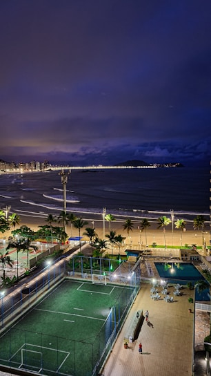 A nighttime coastal scene with a view of a beach, ocean waves, and a long line of lights along the distant shoreline. In the foreground, there is a well-lit sports field with green turf, surrounded by a fence. Adjacent to the field is a swimming pool with lounge chairs nearby. Palm trees line the beach, and a few people can be seen walking on a paved area.