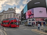 A bustling city scene at a famous intersection featuring a large red double-decker bus in the foreground. Prominent advertising screens display brands like Coca Cola, Gucci, and Samsung. Historic architecture with detailed stonework and a domed structure is visible in the background. Pedestrians and a few vehicles are present on the street.