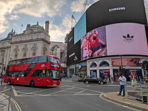 A bustling city scene at a famous intersection featuring a large red double-decker bus in the foreground. Prominent advertising screens display brands like Coca Cola, Gucci, and Samsung. Historic architecture with detailed stonework and a domed structure is visible in the background. Pedestrians and a few vehicles are present on the street.