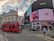 A bustling city scene at a famous intersection featuring a large red double-decker bus in the foreground. Prominent advertising screens display brands like Coca Cola, Gucci, and Samsung. Historic architecture with detailed stonework and a domed structure is visible in the background. Pedestrians and a few vehicles are present on the street.