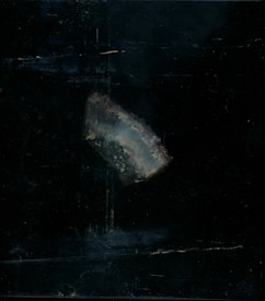 A piece of translucent mineral or crystal with a rough texture is positioned against a dark, almost black background. The mineral appears to have a bluish-gray hue with hints of white and subtle earthy tones.