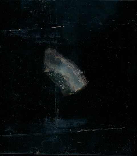 A piece of translucent mineral or crystal with a rough texture is positioned against a dark, almost black background. The mineral appears to have a bluish-gray hue with hints of white and subtle earthy tones.