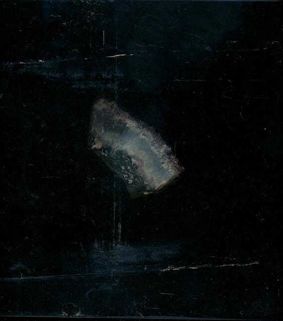 A piece of translucent mineral or crystal with a rough texture is positioned against a dark, almost black background. The mineral appears to have a bluish-gray hue with hints of white and subtle earthy tones.