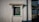 A small rectangular window set in a textured white wall. The window has a green frame and is topped with a decorative wooden canopy. A black drainpipe runs vertically next to the window. To the right, there is a shaded area with a green roof supported by metal framework.
